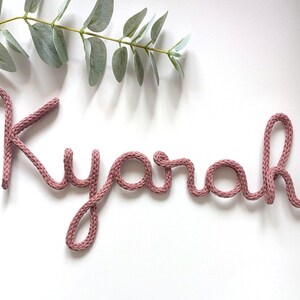 May include: A dusty rose-coloured knitted cord shaped into the cursive name Kyarah. The letters are formed from a single continuous piece of yarn with a visible knit texture, creating a soft, flexible script that spells out the name in lowercase.