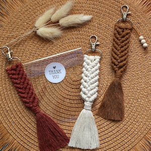 Peut inclure: Trois porte-clés en macramé de couleurs rouge, blanc et marron, chacun avec un pompon et un fermoir en métal. Une pochette en plastique transparent et un autocollant avec les mots "Thank You" sont également visibles. Les porte-clés sont disposés sur une surface tissée de couleur beige.