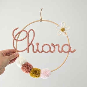 Könnte beinhalten: Ein kreisförmiger Wandbehang aus Holz mit dem Namen Chiara aus altrosa gestricktem Garn. Eine Blume aus weißem und gelbem Garn ist oben rechts befestigt, und vier Pompons in Weiß, Altrosa, Senfgelb und Hellrosa säumen den unteren Bereich.