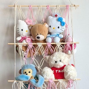 Könnte beinhalten: Ein Makramee-Wandregal mit drei Holzregalen, das verschiedene Plüschtiere enthält. Zu den Spielzeugen gehören eine weiße Katze mit rosa Schleife, ein brauner Bär, eine weiße Katze mit Krankenschwestermütze, ein rosa Schwein, ein blauer Affe und ein weißer Bär mit rotem Hemd.