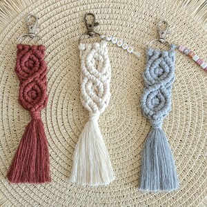 May include: Three macrame keychains with tassels. The keychains are in red, white, and gray. Each keychain has a silver key ring.