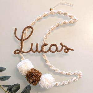 Op de afbeelding: Een wandhanger in de vorm van een halve maan gemaakt van off-white macramékoord met de naam Lucas in bruin koord. Het ontwerp bevat drie pompons in wit en bruin aan de onderkant en een kleine gouden kraal aan de bovenkant.