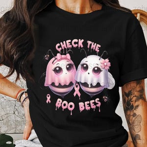 May include: Black t-shirt with a pink and white graphic of two cartoon bees dressed as ghosts, with the text "CHECK THE BOO BEES". The design includes pink ribbons and small spiders, promoting breast cancer awareness.