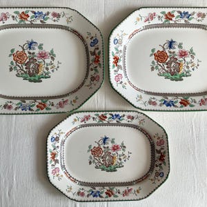 May include: Set of three octagonal serving platters with a floral design. The white platters feature a central bouquet of flowers in shades of orange, pink, and blue. A decorative border with flowers and a green edge complete the design.