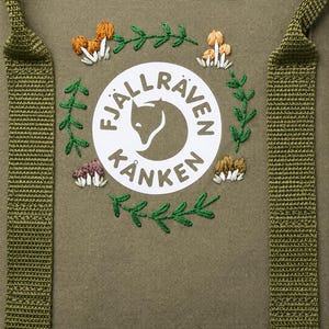 Può includere: Uno zaino verde oliva con una toppa circolare bianca del logo Fjällräven Kånken. Il logo è circondato da dettagli ricamati a mano, tra cui rami con foglie verdi e piccoli gruppi di funghi nei toni dell'arancione, del marrone e del bianco.
