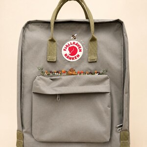 May include: A grey backpack featuring olive green top handles and a front pocket. The front displays a circular Fjällräven Kånken logo, a small embroidered bee, and a row of embroidered colourful mushrooms and greenery along the pocket seam.