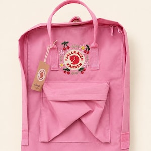 May include: A pink backpack featuring a rectangular shape, two top handles, and a front zippered pocket. The front displays a circular Fjällräven Kånken logo surrounded by embroidered flowers and cherries, with a branded cardboard tag attached.