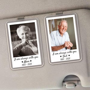 May include: Two rectangular car visor accessories with photos of a woman and a man. Each photo is in a clear frame and has the text "I am always with you" and the names "Mom" and "Dad" with dates. The accessories are clipped to a gray car visor.