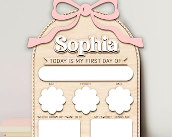 May include: A light-colored wooden milestone board featuring a pink bow at the top, the name Sophia, and the text Today is my first day of. It includes blank spaces for age, height, date, when I grow up I want to be, and my favorite things are.