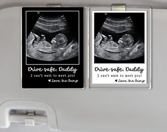 Puede incluir: Dos clips rectangulares para visera de coche, cada uno mostrando una imagen de ultrasonido de un feto. Uno está enmarcado en negro, el otro en blanco. Ambos incluyen el texto "Drive safe, Daddy, I can't wait to meet you! Love, the Bump."