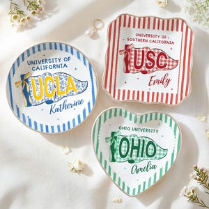 May include: Three decorative ceramic dishes with university logos. A round dish with a blue and yellow UCLA design, a square dish with a red and white USC design, and a heart-shaped dish with a green Ohio University design.