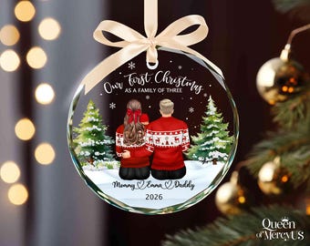 Puede incluir: Un adorno navideño redondo de cristal transparente con una ilustración impresa de una familia de tres personas vista desde atrás. El texto dice Our First Christmas AS A FAMILY OF THREE, Mommy Emma Daddy y 2026. Tiene una cinta color crema.