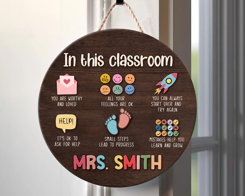 K&ouml;nnte beinhalten: Ein rundes Schild aus braunem Holz mit dem Text "In this classroom" und einer Vielzahl von bunten Illustrationen und Texten. Die Illustrationen beinhalten eine Rakete, ein Herz in einem Umschlag, Fu&szlig;abdr&uuml;cke und eine Vielzahl von Smiley-Gesichtern. Der Text enth&auml;lt S&auml;tze wie "You are worthy and loved", "All your feelings are ok", "You can always start over and try again", "It's ok to ask for help", "Small steps lead to progress", und "Mistakes help you learn and grow". Das Schild enth&auml;lt auch den Text "Mrs. Smith" in gro&szlig;en, bunten Buchstaben.