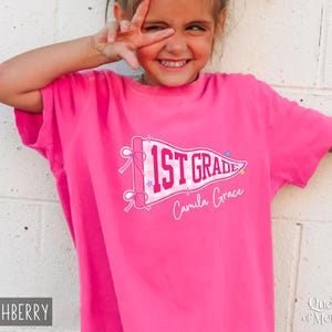 May include: A bright pink short-sleeved t-shirt featuring a white pennant graphic with a pink gingham pattern. The text 1ST GRADE is printed in bold pink letters on the pennant, with the name Camila Grace written below in white script.