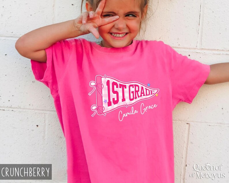 May include: A bright pink short-sleeved t-shirt featuring a white pennant graphic with a pink gingham pattern. The text 1ST GRADE is printed in bold pink letters on the pennant, with the name Camila Grace written below in white script.