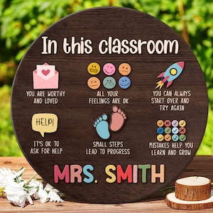 K&ouml;nnte beinhalten: Ein rundes Schild aus Holz mit dem Text "In this classroom" und bunten Illustrationen und Text. Die Illustrationen beinhalten eine Rakete, ein Herz in einem Umschlag, einen Fu&szlig;abdruck und ein Raster aus H&auml;kchen und Kreuzen. Der Text beinhaltet "You are worthy and loved", "All your feelings are ok", "You can always start over and try again", "Help! It's ok to ask for help", "Small steps lead to progress", "Mistakes help you learn and grow", und "Mrs. Smith".