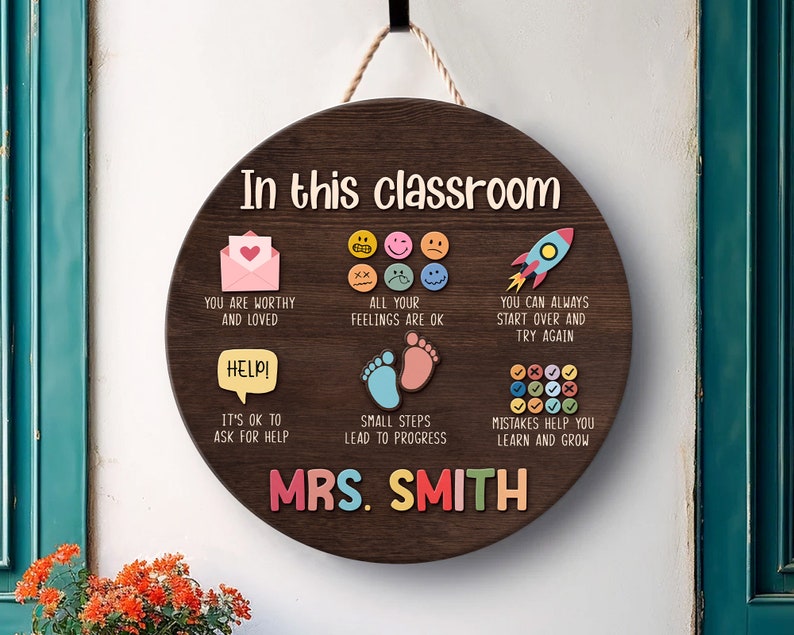 K&ouml;nnte beinhalten: Ein rundes Schild aus braunem Holz mit dem Text "In this classroom" und bunten Illustrationen und Text. Der Text beinhaltet "You are worthy and loved", "All your feelings are ok", "You can always start over and try again", "It's ok to ask for help", "Small steps lead to progress", "Mistakes help you learn and grow", und "Mrs. Smith".