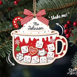 May include: A wooden Christmas ornament shaped like a mug filled with hot cocoa, featuring a red and white gingham bow, a candy cane, and white whipped cream labeled "The Johnson". The mug contains marshmallow-shaped charms with names.