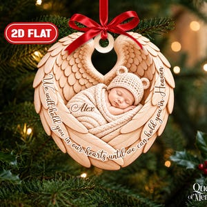 Puede incluir: Un adorno navideño de madera plano en 2D que muestra a un bebé durmiendo envuelto en una manta, centrado dentro de alas de ángel talladas. El texto dice "We will hold you in our hearts until we can hold you in heaven" con el nombre "Alex".