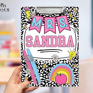 May include: A rectangular clipboard featuring a black and white leopard print background. The design includes pink banners with the text MRS. SANDRA, a yellow lightbulb, a pink apple, a rainbow, daisies, and a lightning bolt graphic.