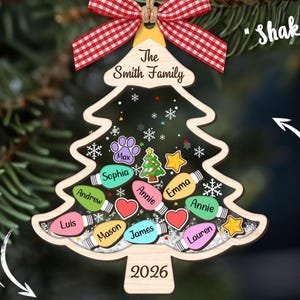 Puede incluir: Un adorno navideño de madera con forma de árbol, un lazo de cuadros rojo y blanco, el texto The Smith Family y 2026. El centro contiene formas de bombillas de colores con nombres, una huella, un corazón y una estrella sobre purpurina blanca.