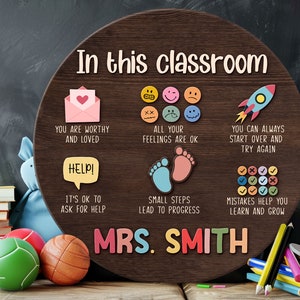 K&ouml;nnte beinhalten: Ein rundes Schild aus braunem Holz mit dem Text "In diesem Klassenzimmer" und verschiedenen positiven Botschaften f&uuml;r Sch&uuml;ler. Das Schild enth&auml;lt auch den Namen "MRS. SMITH" in bunten Buchstaben.