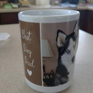 Puede incluir: Taza de cer&aacute;mica blanca con un panel marr&oacute;n con el texto "Best Dog Dad" y un coraz&oacute;n blanco. El otro lado de la taza muestra un perro husky blanco y negro con ojos azules. Una l&aacute;mpara es visible en el fondo.