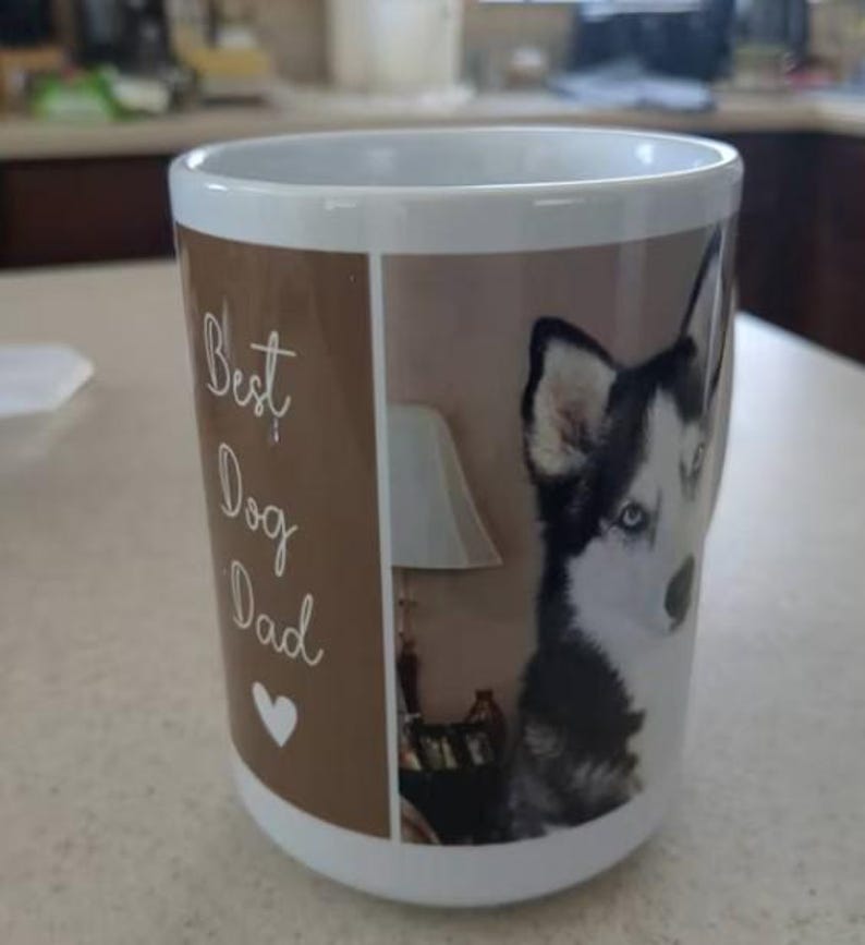 Puede incluir: Taza de cer&aacute;mica blanca con un panel marr&oacute;n con el texto "Best Dog Dad" y un coraz&oacute;n blanco. El otro lado de la taza muestra un perro husky blanco y negro con ojos azules. Una l&aacute;mpara es visible en el fondo.