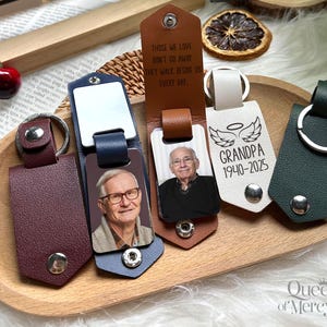 May include: A collection of personalized leather keychains in various colors, including burgundy, navy, and green. Some keychains feature photos of a person, while others have the text "GRANDPA 1940-2025" and the quote "THOSE WE LOVE DON'T GO AWAY THEY WALK BESIDE US EVERY DAY."