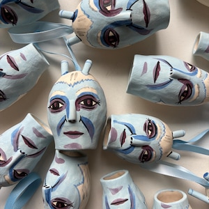 May include: A group of light blue ceramic face ornaments with painted purple, white, and beige features. Each ornament has a light blue ribbon loop for hanging. The faces have a whimsical, artistic style.