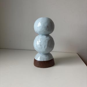 May include: A ceramic sculpture with a stacked design. The top three spheres are a light blue color, and the base is brown. The sculpture is on a white surface.