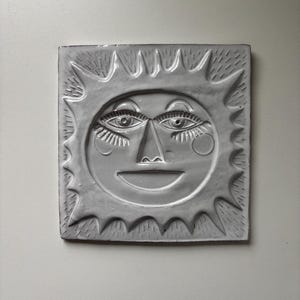 Może przedstawiać: Kwadratowa, szara płytka ceramiczna przedstawiająca uśmiechniętą twarz słońca ze szczegółowymi oczami, nosem i ustami. Słońce ma promienie wokół twarzy. Płytka to element dekoracyjny, być może na ścianę lub jako podstawka.