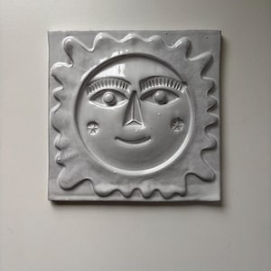 May include: A square, grey ceramic tile depicting a smiling sun face with detailed eyes, nose, and mouth. The sun has a wavy, textured border and star-like accents on each cheek. The tile is mounted on a white wall.