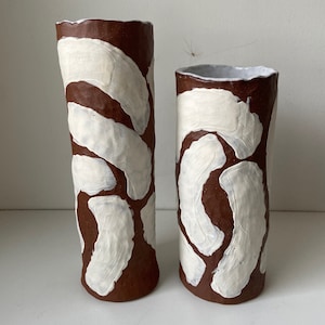 Puede incluir: Dos jarrones de cerámica con base marrón y diseños abstractos blancos. Los jarrones son cilíndricos y tienen una superficie texturizada.