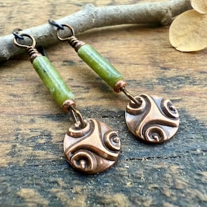 May include: A pair of handcrafted earrings featuring green cylindrical beads and copper-colored circular pendants with a spiral design. The earrings are suspended from black ear wires and are displayed on a wooden surface.