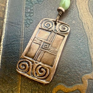 May include: A copper-colored pendant with an Art Deco design, featuring a rectangular shape with spiral and geometric details. The pendant is suspended from a green bead and a copper chain. The pendant rests on a textured, dark background.
