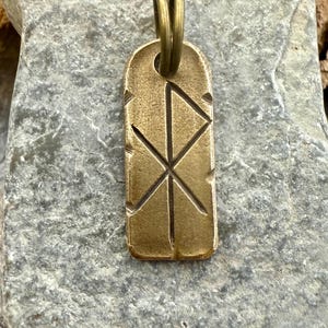 May include: A rectangular brass pendant with a carved geometric design and a double loop bail. The pendant has a worn, antique appearance. The design features intersecting lines forming an X and a triangle. The pendant is displayed on a gray stone surface.