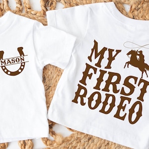 May include: Two white t-shirts with brown graphics. One shirt features a horseshoe with the name "Mason" inside. The other shirt says "My First Rodeo" with a cowboy riding a horse. Both shirts are short-sleeved.