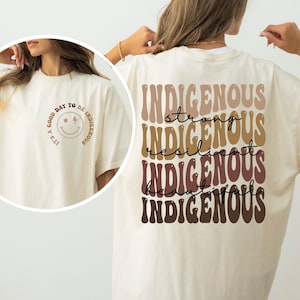 May include: Cream-colored t-shirt with the text "Indigenous" stacked vertically in a retro font, with the words "strong, resilient, beautiful" in between. The front of the shirt has a smiley face with the text "It's a good day to be Indigenous."