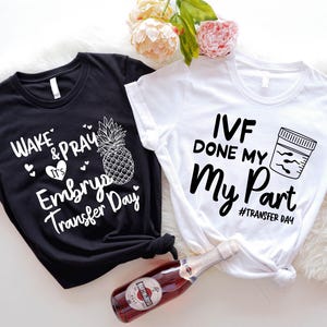 May include: Two t-shirts, one black and one white, with text and graphics. The black shirt says "Wake & Pray It's Embryo Transfer Day" with a pineapple graphic. The white shirt says "IVF Done My Part" with a sperm sample graphic.