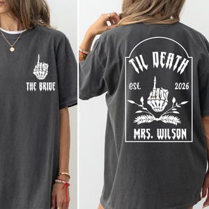 May include: Dark gray t-shirt with white graphic text and design. The front of the shirt says "THE BRIDE" with a skeleton hand giving the middle finger. The back says "TIL DEATH est. 2026 MRS. WILSON".