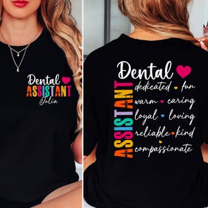 Puede incluir: Camiseta negra con la frase "Dental Assistant" en colores vibrantes, un corazón rosa y el nombre "Julia". La parte trasera de la camiseta enumera cualidades como "dedicado", "divertido" y "compasivo".