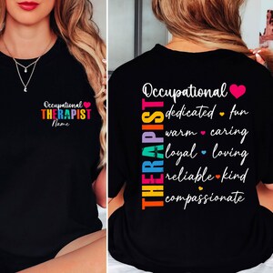 Puede incluir: Camiseta negra con las palabras "Occupational Therapist" en texto colorido en la parte delantera. La parte posterior de la camiseta presenta una lista de atributos positivos como "dedicado", "atento" y "compasivo".