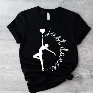 May include: Black t-shirt featuring a white silhouette of a dancer holding a heart-shaped balloon. The words "just dance" are written in a cursive font around the dancer. The shirt is knotted at the bottom.