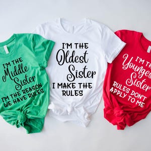 Puede incluir: Tres camisetas de manga corta en verde, blanco y rojo. La camiseta verde dice I'M THE MIDDLE SISTER I'M THE REASON WE HAVE RULES. La blanca dice I'M THE OLDEST SISTER I MAKE THE RULES. La roja dice I'M THE YOUNGEST SISTER RULES DON'T APPLY TO ME.