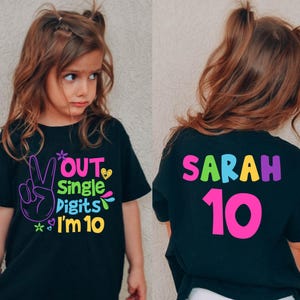Könnte beinhalten: Schwarzes T-Shirt mit farbenfrohem Text und Grafiken. Auf der Vorderseite steht "Out Single Digits I'm 10" mit einem Friedenszeichen. Auf der Rückseite steht "Sarah 10" in leuchtenden Farben. Das T-Shirt ist für ein Kind.