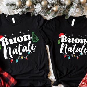 May include: Two black t-shirts featuring the white text Buon Natale, a red Santa hat, a green Christmas tree, and a string of colourful holiday lights. The shirts are surrounded by red and black plaid fabric and festive greenery.