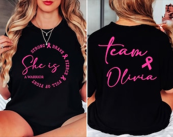 May include: Black t-shirt with pink text and a pink ribbon. The front of the shirt says "She is a Warrior" inside a circle with "Strong, Brave, Fierce, Full of Fight". The back says "Team Olivia".