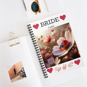 May include: A spiral bound notebook with a white cover featuring a pink and white flower with a ladybug on it. The cover also has the text "BRIDE crew" with red hearts.