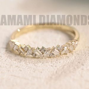 May include: A gold band ring featuring a line of small, sparkling diamonds. The diamonds are set in various shapes, including square and baguette cuts. The ring rests on a light-coloured surface. The text "AMAMI DIAMONDS" is visible.